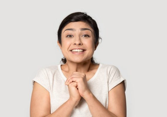 Head shot portrait close up smiling Indian girl joining hands with excitement, surprised beautiful young woman looking at camera, excited by good news, isolated on grey studio background